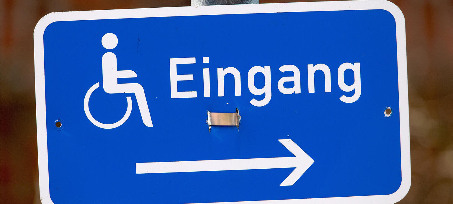  ein kleines blaues, schief hängendes Hinweisschild mit einem Rollstuhl-Piktogramm, der Aufschrift 'Eingang' und einem Pfeil nach rechts an einem Schildermast - BGG